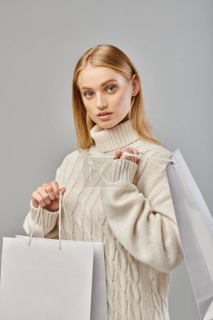 trendy blonde woman in warm knitted sweater with white shopping bags looking at camera on grey