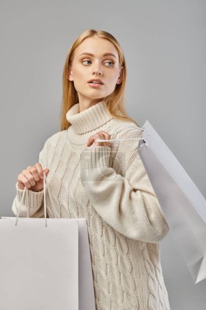 stylische blonde Frau im weißen Strickpullover mit Einkaufstaschen, die wegschauen auf grau, Winterschlussverkauf