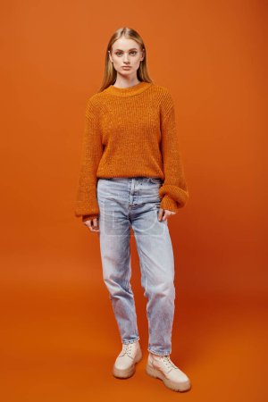 Téléchargez les photos : Longueur totale de femme blonde élégante en pull tricoté et jean bleu sur fond orange, hiver - en image libre de droit