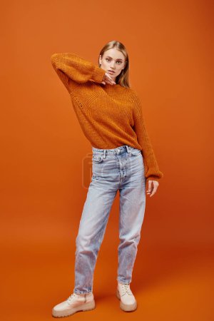 Téléchargez les photos : Femme blonde tendance en pull tricoté chaud et jean bleu sur fond orange, mode d'hiver - en image libre de droit