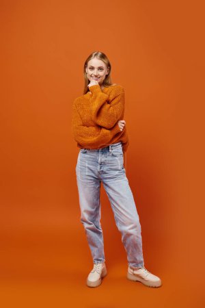 Téléchargez les photos : Joyeuse femme blonde en pull d'hiver orange et jean bleu souriant à la caméra sur fond lumineux - en image libre de droit