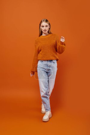 Téléchargez les photos : Femme blonde tendance en jeans bleus et pull d'hiver orange marchant sur fond lumineux, pleine longueur - en image libre de droit