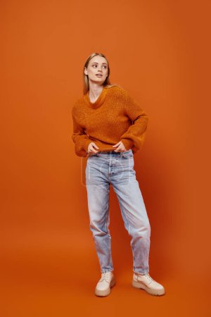Téléchargez les photos : Pleine longueur de femme blonde élégante en jeans bleus et pull d'hiver regardant loin sur fond orange - en image libre de droit
