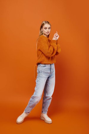 Téléchargez les photos : Pleine longueur de charmante femme blonde en jeans bleus et pull chaud sur fond orange, style hiver - en image libre de droit