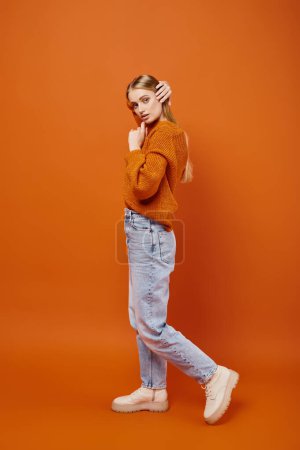 Téléchargez les photos : Séduisante femme blonde en jeans et pull tricoté chaud posant sur fond orange, tendance hivernale - en image libre de droit
