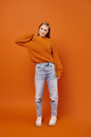 Téléchargez les photos : Pleine longueur de femme blonde à la mode en jeans et pull tricoté doux posant sur fond orange - en image libre de droit