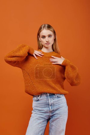 Téléchargez les photos : Femme blonde expressive en chandail tricoté lumineux et jeans bleus sur fond orange, mode moderne - en image libre de droit
