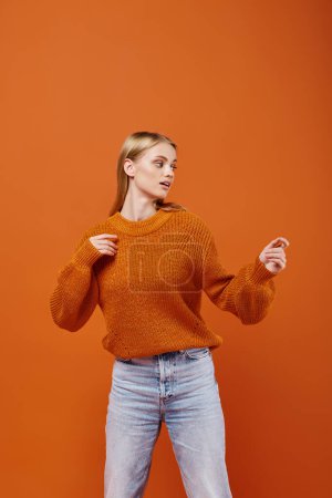 Téléchargez les photos : Femme blonde sensuelle en pull lumineux et jeans bleus regardant loin sur fond orange, style d'hiver - en image libre de droit