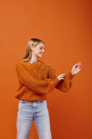 Téléchargez les photos : Femme blonde heureuse regardant son pull tricoté chaud et lumineux sur fond orange, hiver confortable - en image libre de droit