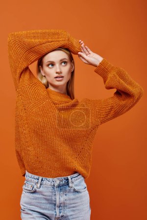 Téléchargez les photos : Jeune femme blonde en chandail tricoté orange posant avec les bras au-dessus de la tête sur orange, vibes d'hiver - en image libre de droit