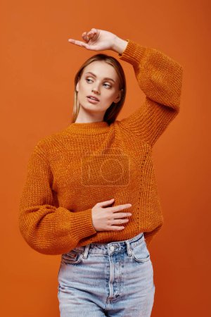blonde woman in bright winter sweater posing with hand above head pointing with finger on orange