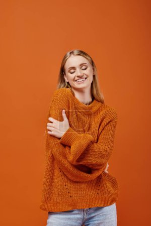pleased woman in warm knitted sweater hugging herself and smiling with closed eyes on orange