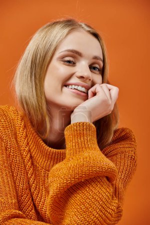 Porträt einer fröhlichen blonden Frau in buntem Strickpullover mit natürlichem Make-up auf orangefarbenem Hintergrund