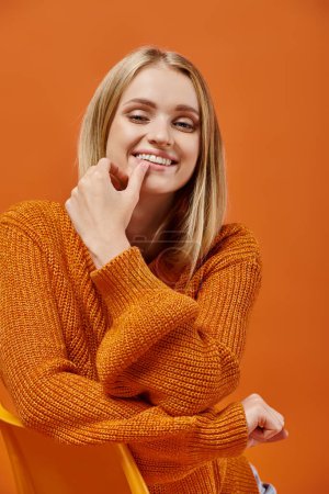 portrait of joyful blonde woman in bright and warm knitted sweater with natural makeup on orange