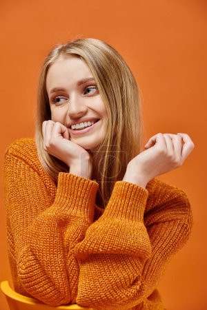 smiley blonde woman in bright and cozy knitted with natural makeup looking at camera on orange