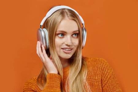 positive blonde Frau in hellem Winterpullover und Kopfhörer, die Musik auf orangefarbenem Hintergrund hört