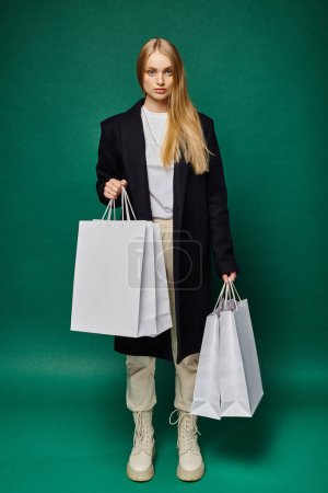 full length of trendy blonde woman in black coat with white shopping bags on green, seasonal sales