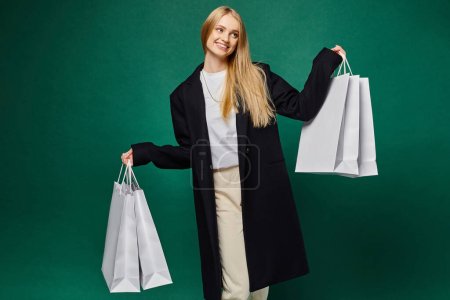 Téléchargez les photos : Femme blonde souriante en manteau noir tendance tenant des sacs à provisions blancs sur vert, joie d'hiver - en image libre de droit