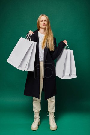 trendy blonde woman in black coat holding white shopping bags and pouting lips on green, full length