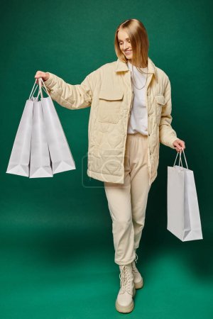 full length of cheerful blonde woman in trendy winter attire holding white shopping bags on green