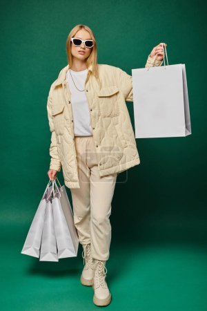 Téléchargez les photos : Femme blonde à la mode en vêtements d'hiver et lunettes de soleil posant avec des sacs à provisions sur fond vert - en image libre de droit