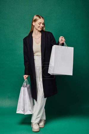 full length of smiling blonde woman in warm black coat looking at white shopping  bags on green