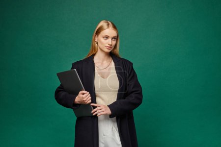 Photo for Trendy blonde woman in black winter coat holding laptop and looking away on green backdrop - Royalty Free Image