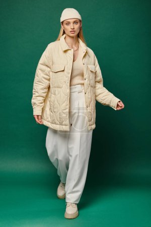 Charmantes und trendiges weibliches Modell in elfenbeinfarbener Jacke und Mütze, das auf grüner Wintermode wandelt