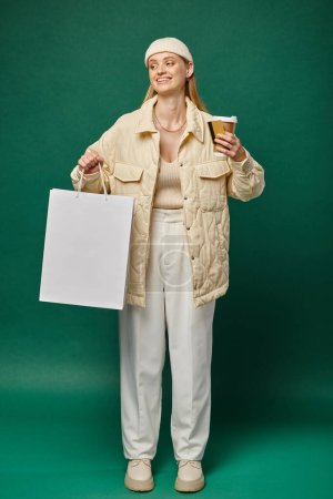 happy woman in stylish winter clothing with shopping bag, takeaway drink and credit card on green