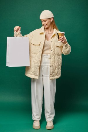 cheerful woman in trendy winter attire with shopping bag, hot drink and credit card on green