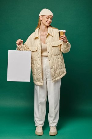 fröhliche junge Frau in trendiger Winterkleidung mit Einkaufstasche, Heißgetränk und Kreditkarte auf grün