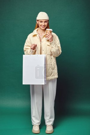 Téléchargez les photos : Heureux jeune femme en tenue d'hiver élégant avec sac à provisions et boisson chaude dans une tasse en papier sur vert - en image libre de droit