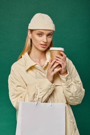 charming woman in warm jacket and beanie hat with shopping bag and paper cup on green, winter style