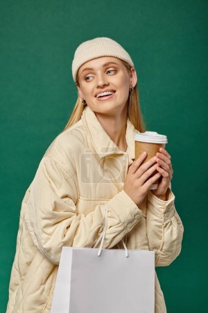 Téléchargez les photos : Femme insouciante en veste d'hiver à la mode et bonnet chapeau avec sac à provisions et boisson chaude sur vert - en image libre de droit