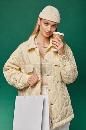 stylische Frau in kuscheliger Winterkleidung mit Einkaufstasche und Imbissgetränk blickt in die Kamera auf Grün
