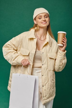 joyful woman in warm winter clothes with shopping bag and paper cup looking at camera on green