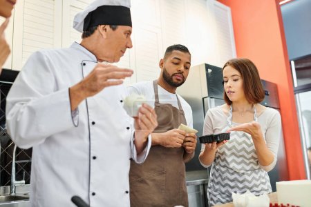Téléchargez les photos : Beaux jeunes gens diversifiés avec tabliers consultation avec leur chef pendant les cours de cuisine - en image libre de droit