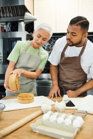 mature beautiful woman and african american man with aprons baking delicious cake, cooking courses