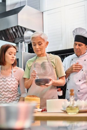 mature woman in apron learning how to use silicone brush on cake next to her young friend and chef