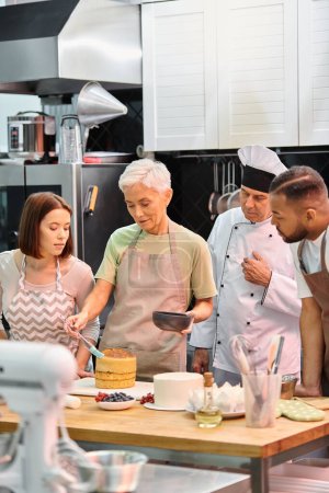 mature woman in apron brushing cake with syrup on cake next to diverse friends and chef in white hat
