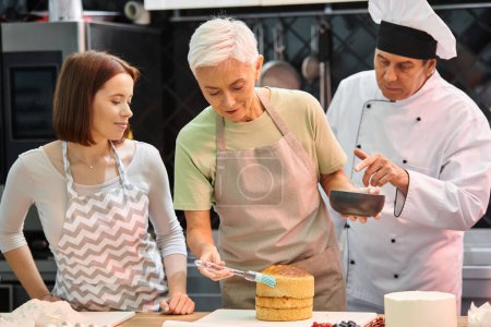 mature woman in apron brushing cake with syrup on cake next to young friend and chef in white hat