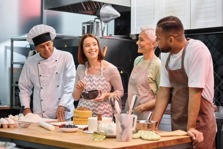 Photo for Cheerful young woman brushing cake with syrup and smiling at her diverse friends next to mature chef - Royalty Free Image