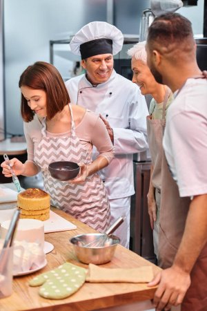 joven alegre mujer cepillando pastel con jarabe mientras sus diversos amigos y chef hablar activamente
