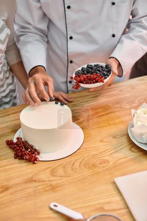 abgeschnittene Ansicht von reifen Koch lehrt seine junge Studentin, wie man Kuchen mit Beeren im Unterricht dekoriert