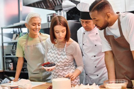 young woman with apron decorating cake next to her multiracial jolly friends and chef in white hat