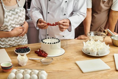 vista recortada de chef maduro decorando delicioso pastel con grosella roja junto a sus diversos estudiantes