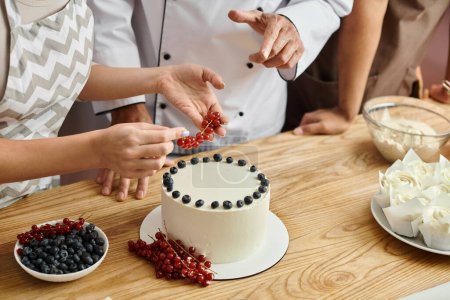 vista recortada de la joven mujer decorando pastel con grosella roja cerca de chef y amigo afroamericano