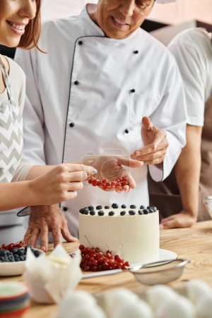 cropped view of jolly young woman decorating cake near mature chef and her african american friend