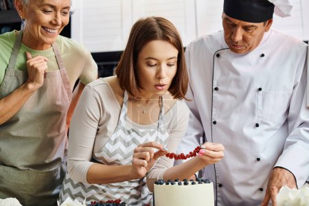 junge Frau mit Schürze dekoriert Kuchen mit roter Johannisbeere neben ihrem fröhlichen reifen Freund und Koch