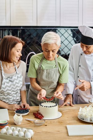 reife Frau in Schürze dekoriert Kuchen mit roter Johannisbeere neben ihrem lustigen jungen Freund und Koch